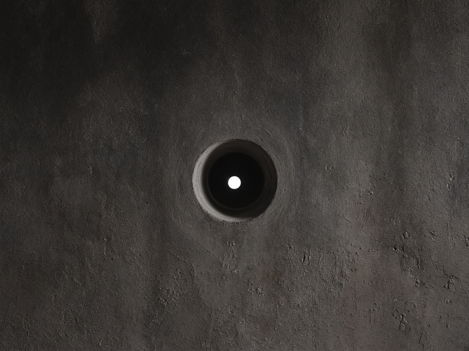 A small circular opening in a dark plaster wall, a point of daylight through it.